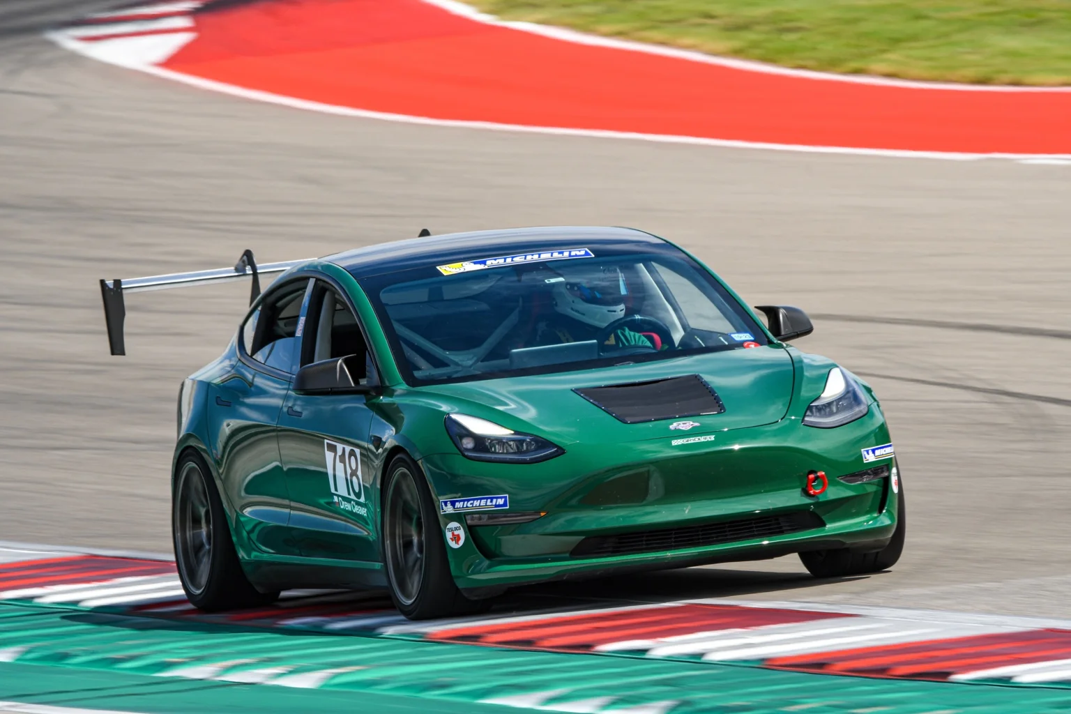 Green Spec Tesla Model 3, number 718, with a rear wing taking the kerb at Circuit of the Americas