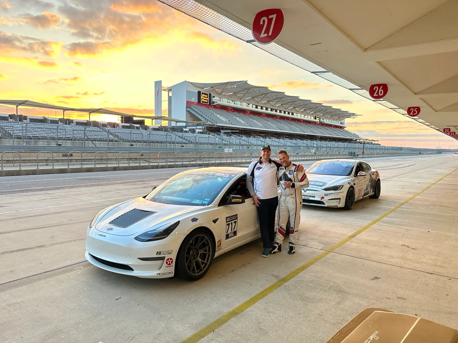 Two white Tesla race cars and two people in the COTA pit lane at sunset